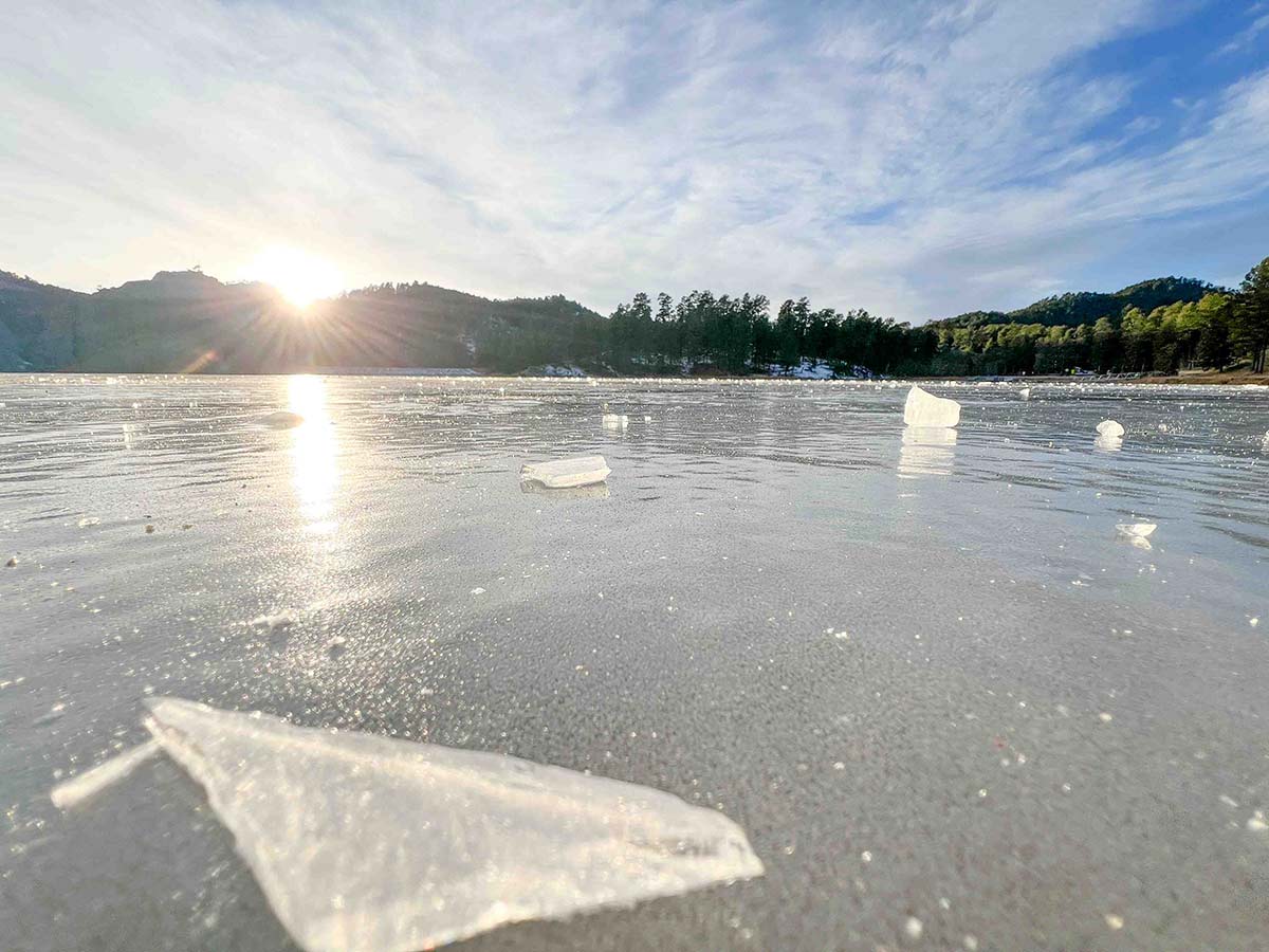 ice-chunks-dot-the-sandy-shoreline-as-golden-sunrise-light-reflects-off-wet-beach.-forested-hills-frame-this-dramatic-winter-coastal-scene-with-streaking-clouds-overhead.