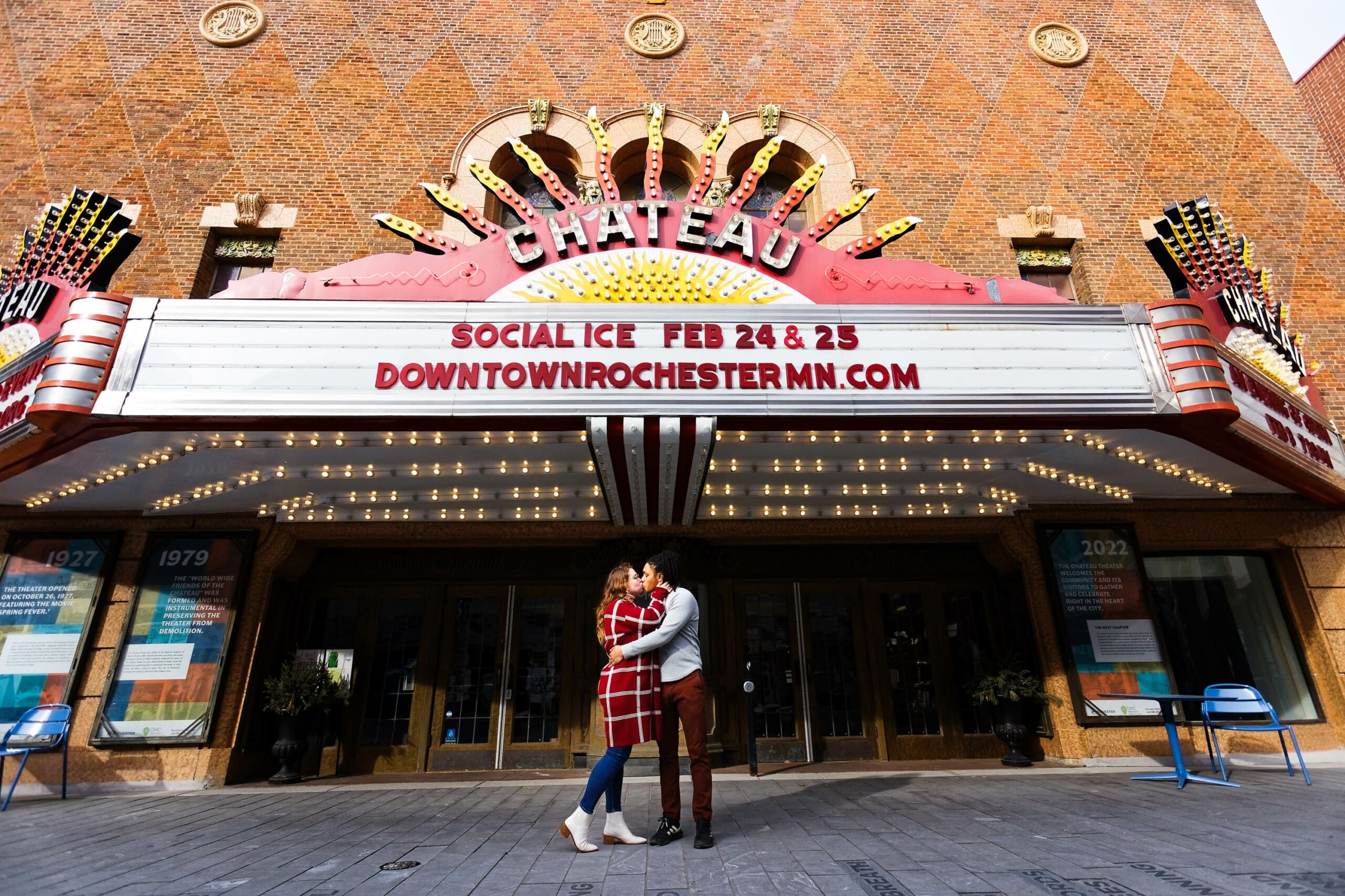 GPT A couple embraces in front of the Chateau Theater's iconic marquee in Rochester, MN. The marquee announces an event, "Social Ice Feb 24 & 25," and the theater's website. The theater's facade is a classic brick design with decorative elements, and the marquee is lit up, suggesting the photo was taken during the day. The couple, dressed in winter clothing, appears to be sharing a joyful moment, capturing a blend of urban architecture and personal celebration.