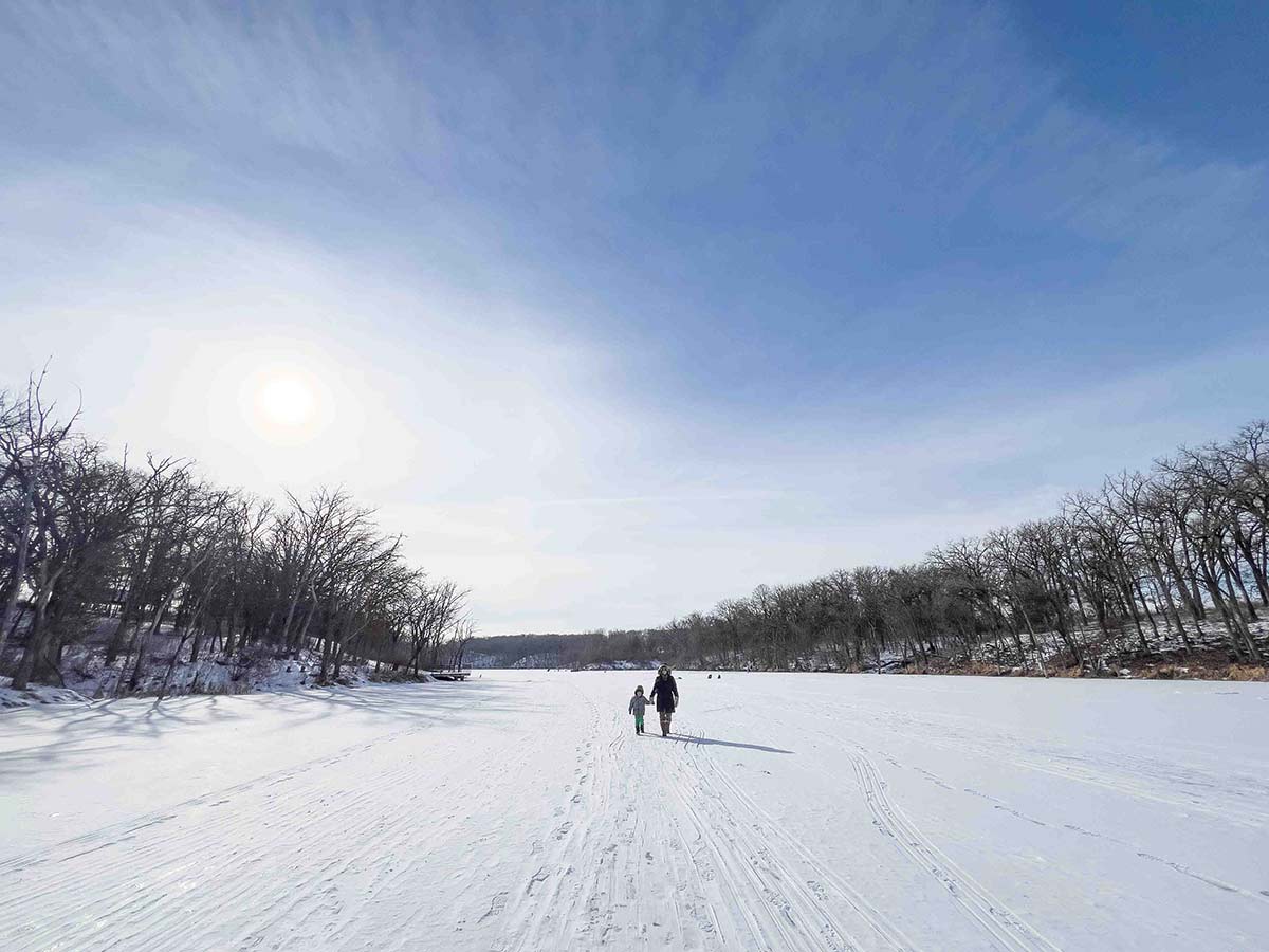 winter-engagement-session-on-frozen-lake-with-couple-walking-hand-in-hand-across-snow-covered-ice-surrounded-by-leafless-trees-under-cloudy-sky