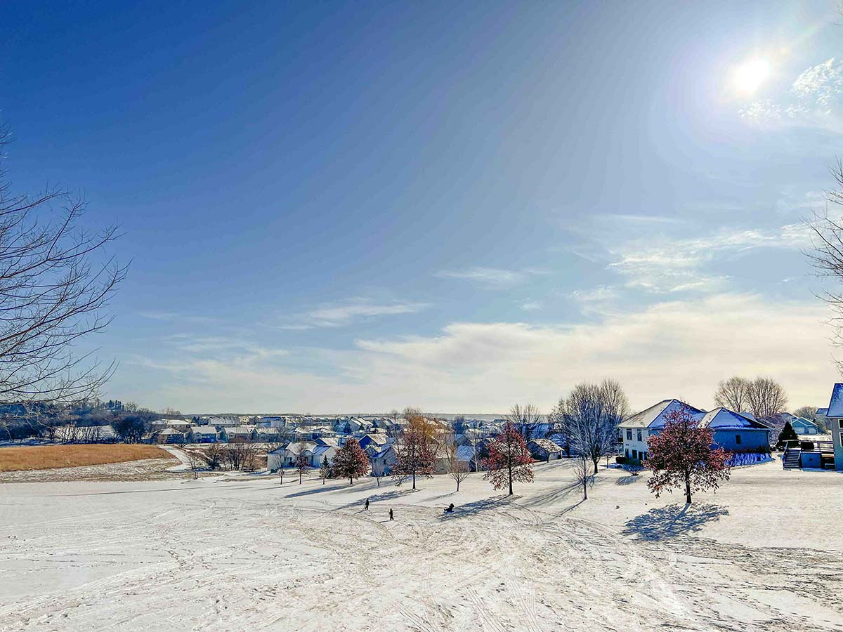 winter-sledding-hill-covered-in-fresh-snow-with-curved-tracks-leading-down-toward-suburban-homes-under-brilliant-blue-sky-and-sunshine