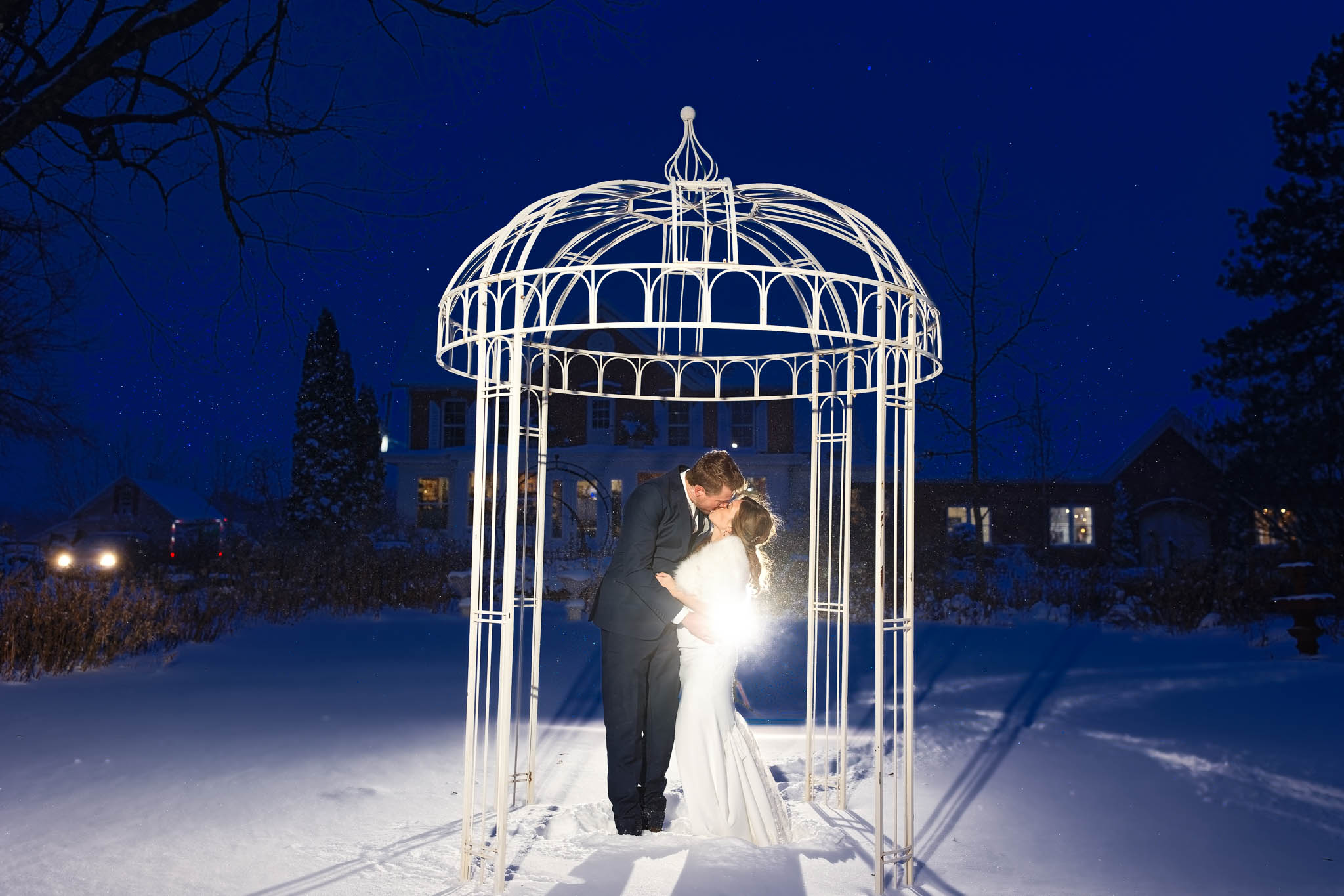 bride and groom winter photos with snow blue hour under pergolla with string light with garten marketplatz in background kissing under ornamental arch dome