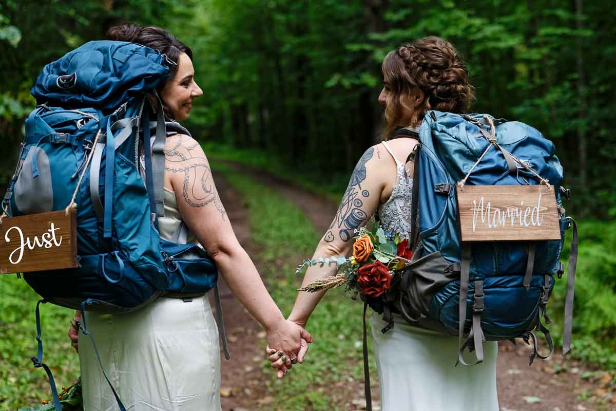 two brides hiking with backpacks and just married sign, outdoor wedding photography in minnesota state park forest trail