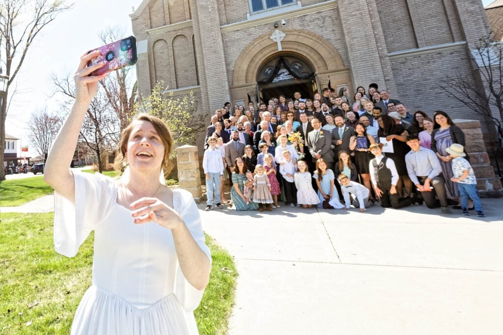Bride snapping a selfie in front of the church, guests gathered and smiling behind her