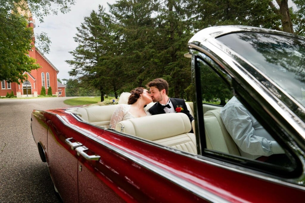 bride and groom seated at the back a of red convertible car kissing